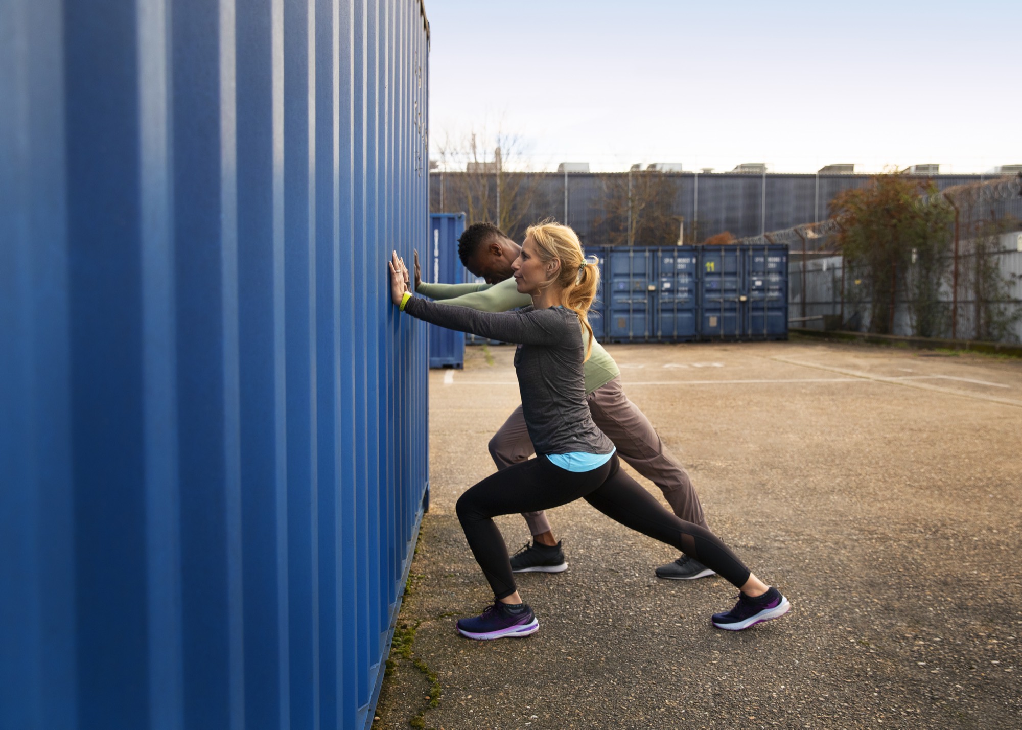 Two people stretching outdoors against a blue container